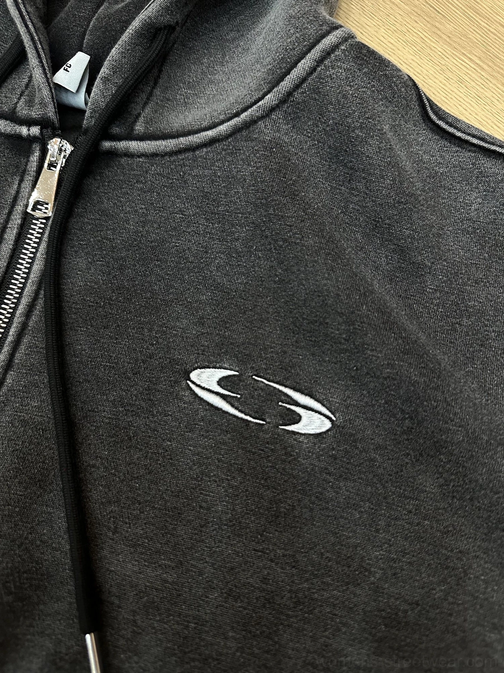 Close-up detail of the dark gray washed cropped hoodie featuring a silver metal zipper, black hood drawstrings, and oval chest embroidery