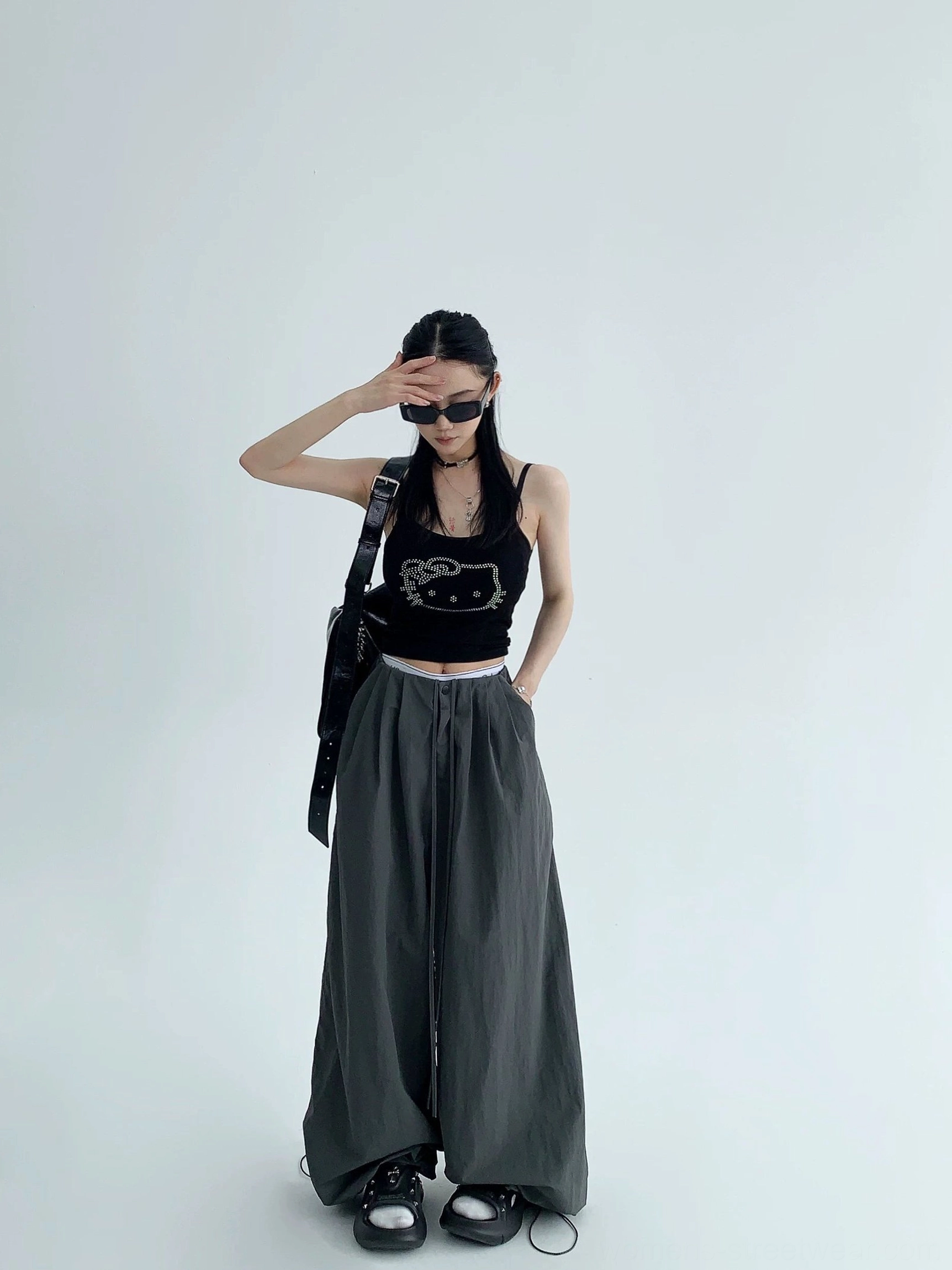 Dark gray wide-leg streetwear parachute pants shown in a full-body front view with black camisole and oval sunglasses