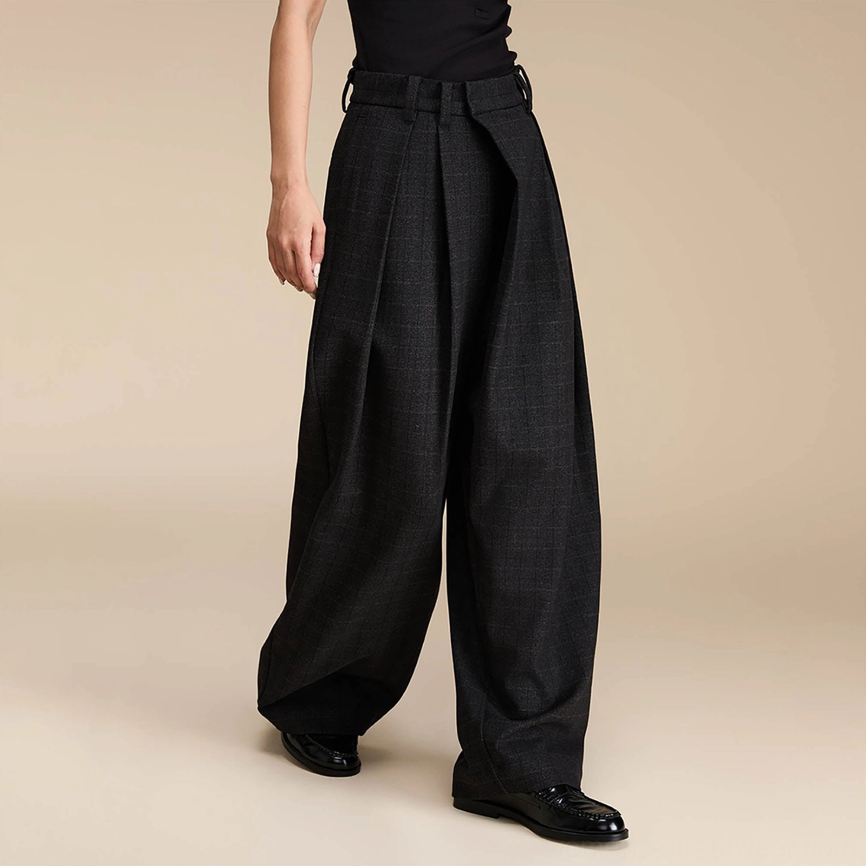 Three-quarter view of dark grey plaid wide-leg streetwear trousers with front pleats, tailored waistband, black top, and leather loafers
