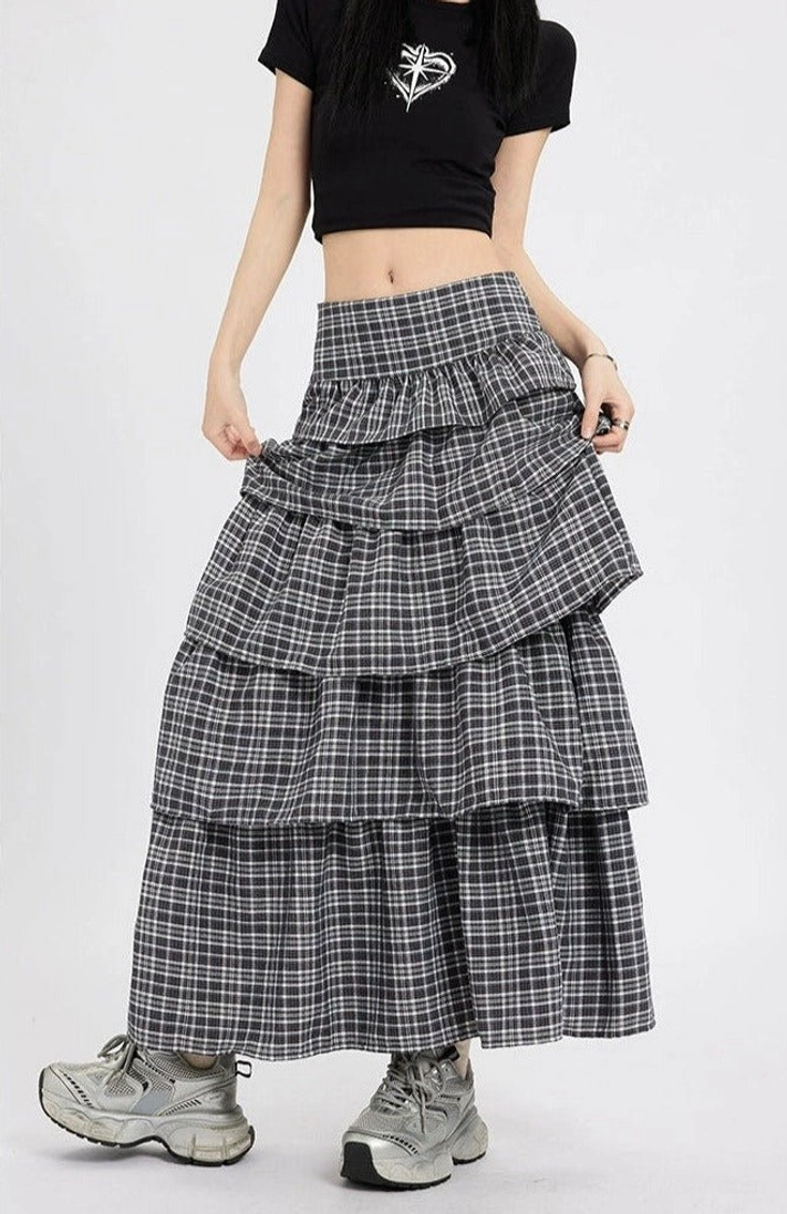 Front three quarter view of a black dark grey plaid tiered maxi skirt with high waist layered ruffles styled with a black cropped tee and silver chunky sneakers