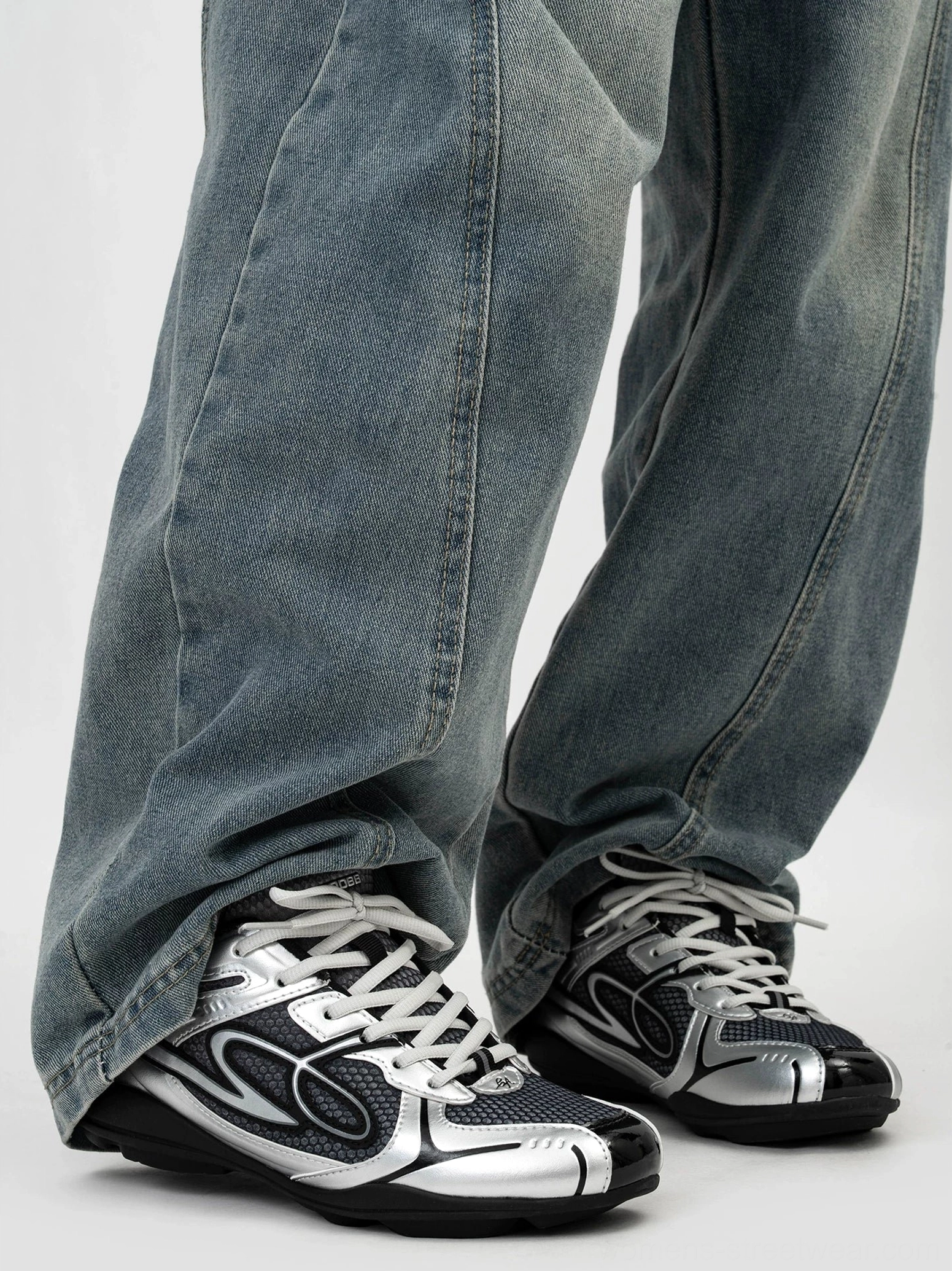 Dark silver low-top retro runner sneaker worn with loose blue denim showing black mesh upper silver overlays and streamlined futuristic streetwear shape