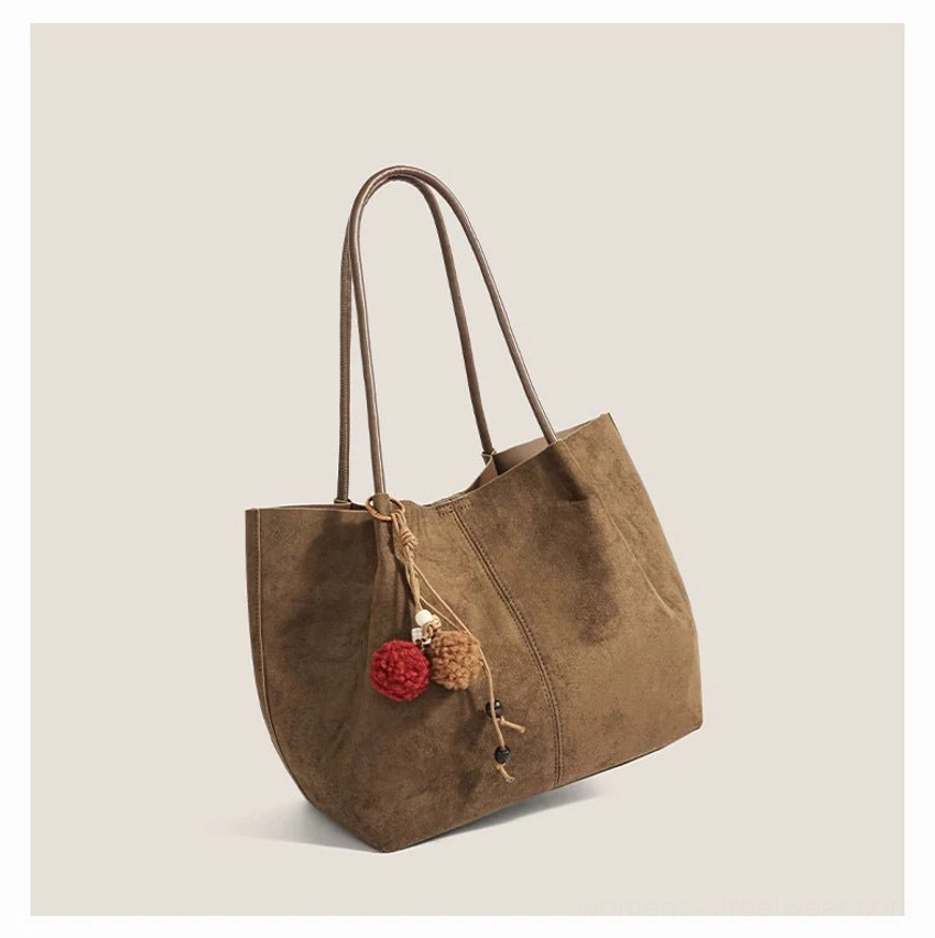 Dark taupe brown faux suede pom charm tote bag shown from a side three quarter perspective on a neutral studio background