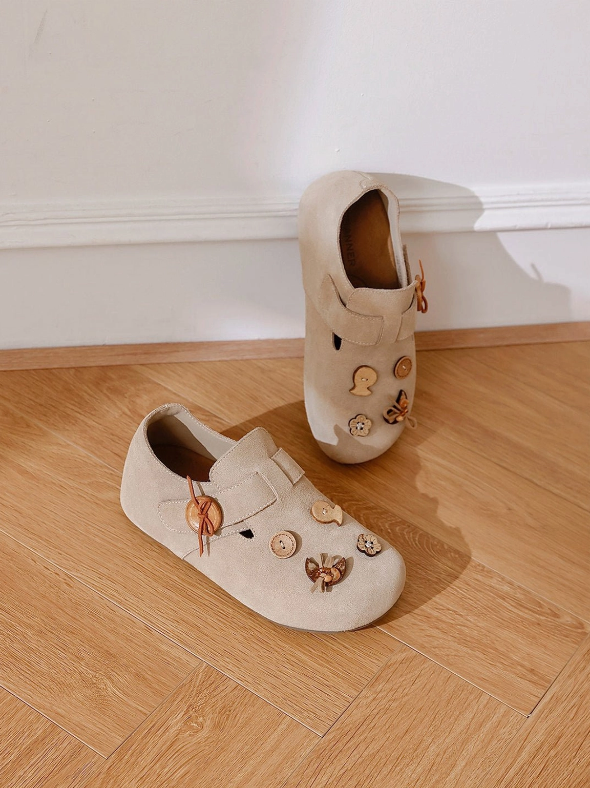 Two pairs of decorative suede-look button strap slip-on shoes displayed on a wooden floor