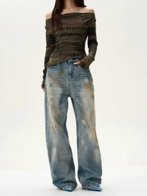 Female model wearing distressed blue wide-leg baggy jeans with vintage washed denim, brown distressed details, and a relaxed Y2K streetwear silhouette