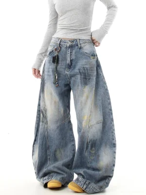 Front three-quarter view of oversized distressed blue wide leg jeans with vintage bleach wash paint splash fading and decorative beaded waist chain styled with a fitted gray top