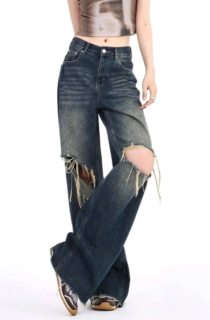 Front cross-legged view of distressed dark wash wide leg ripped jeans with double frayed knee openings, loose high-rise silhouette, gradient denim finish, and vintage footwear