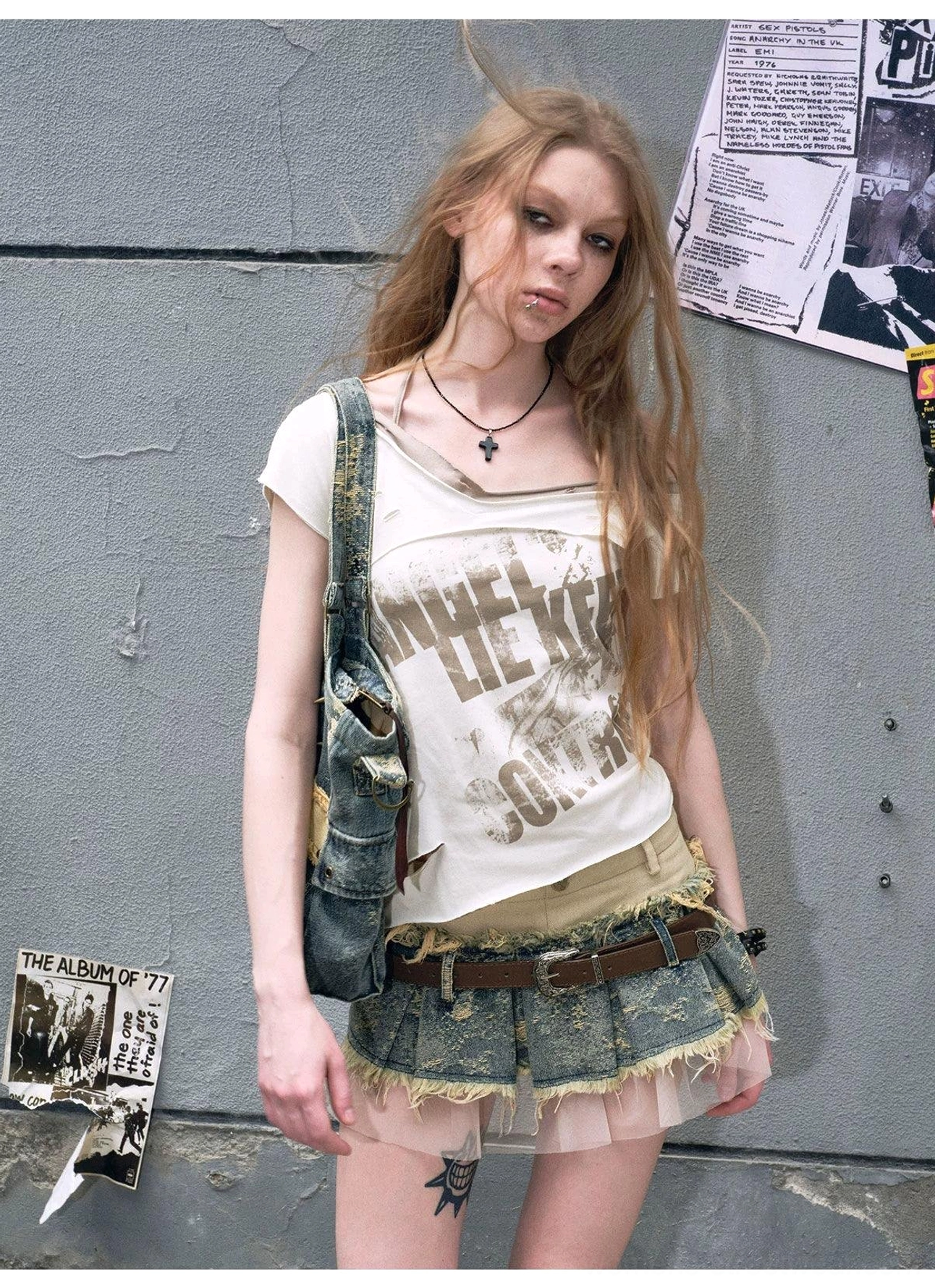 Back three-quarter view of a distressed denim mini skirt with frayed edges, layered tulle hem, brown belt, graphic tee, and denim crossbody bag