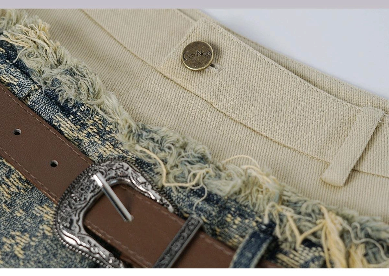 Close-up of the distressed denim mini skirt waistband with vintage engraved metal buckle, brown belt, metal button, and raw denim edge