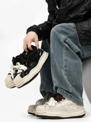 Black distressed double-lace shell-toe chunky streetwear sneakers held at a low angle with a melted-effect platform sole, textured upper, and layered black and cream laces