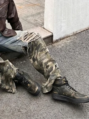 Black distressed leather padded-cuff combat boots styled with camouflage pants and a brown leather jacket in an urban streetwear outfit