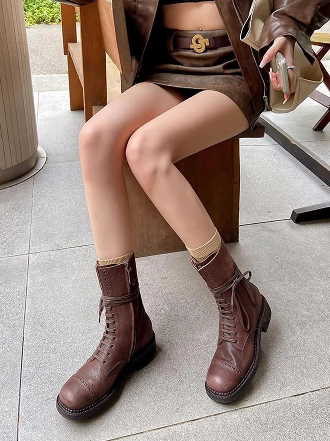 Dark brown distressed leather mid-calf combat boots worn with beige crew socks and brown leather outfit at outdoor cafe
