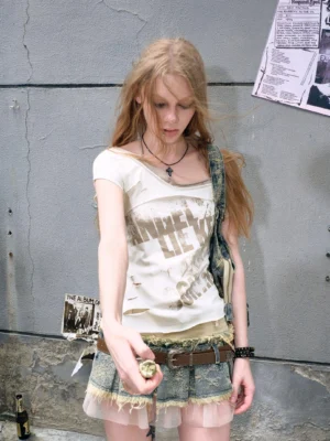 Beige distressed off-shoulder graphic tie-back top worn by a woman with a frayed denim mini skirt against punk poster walls