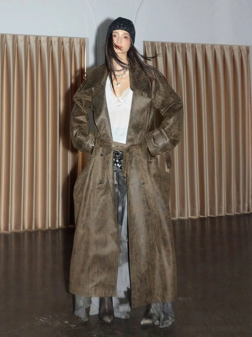 Female model wearing a distressed olive green trench coat with wide lapels, oversized patch pockets, knit beanie, white top, sheer skirt, and boots in a vintage streetwear studio setting