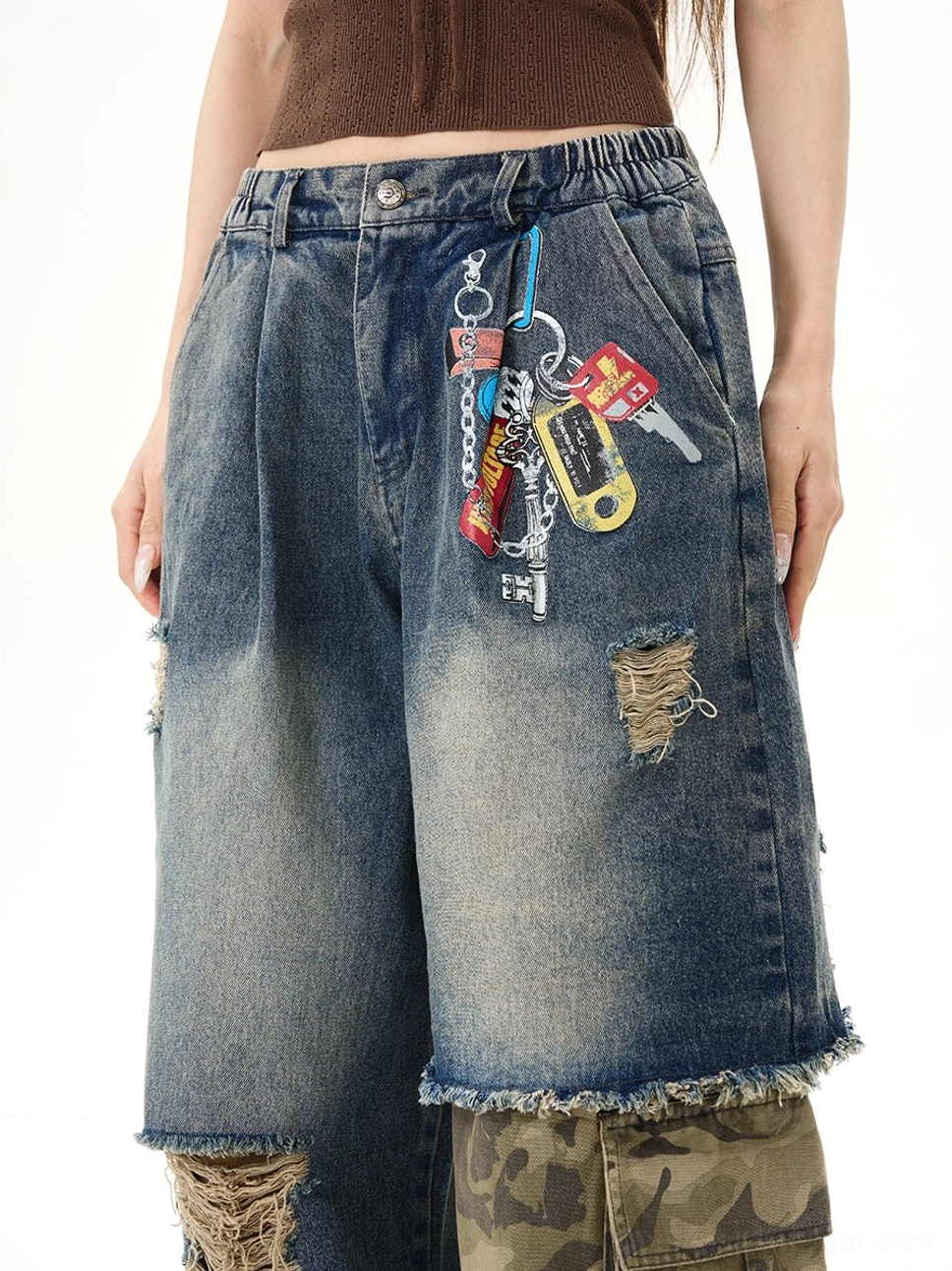 Close-up of distressed patchwork denim jeans waist and thigh area, showing vintage washed blue fabric, front pocket keychain-style embroidery, camo inner patchwork, and raw frayed details