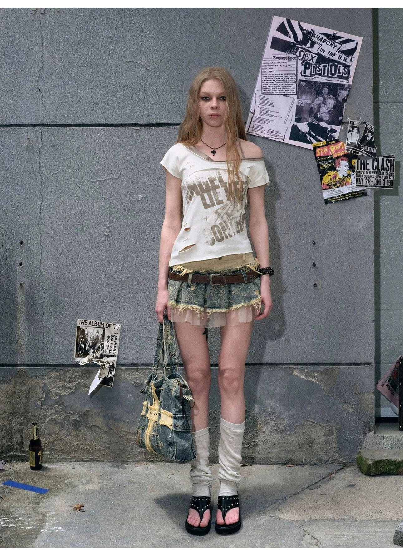 Front view of a distressed patchwork denim mini skirt with sheer layered hem, matching brown waist belt, graphic tee, and denim tote bag