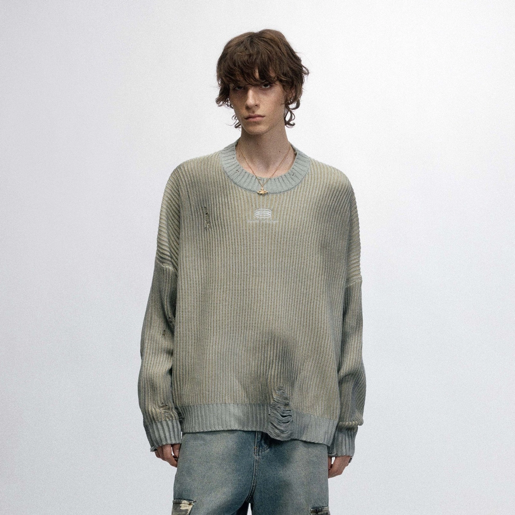 Front view of a male model wearing a sage gray distressed ribbed logo sweater with ripped denim jeans
