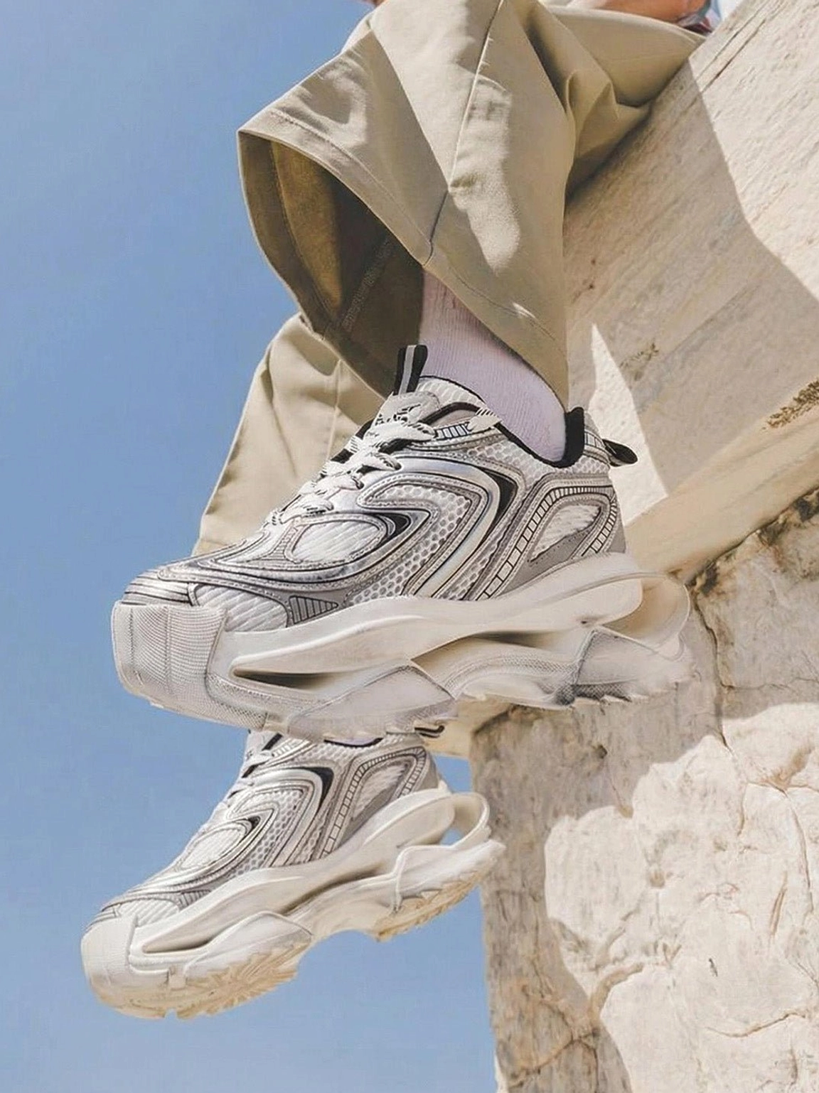 Distressed silver and black dad sneakers with thick sculpted sole styled with khaki cargo pants against a stone wall