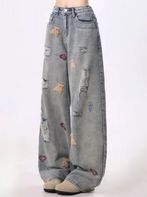 Front low angle view of distressed teddy bear embroidered wide-leg jeans with vintage washed blue denim, red heart details, frayed rips, high waist, and belt loops