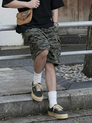 Retro distressed low-top casual sneaker on male model wearing streetwear outfit, three-quarter eye-level view outdoors on street curb, paired with white crew socks and camo shorts