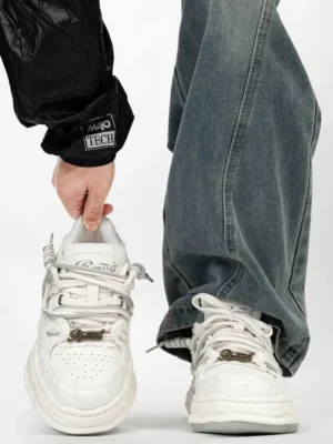 White distressed low-top platform sneakers with layered laces, decorative metal lace charm, weathered upper and chunky off-white sole styled with baggy blue jeans and black streetwear outerwear
