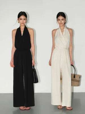 Two women wearing draped halter wide-leg jumpsuits in black and apricot with wrap necklines and flowing full-length pants