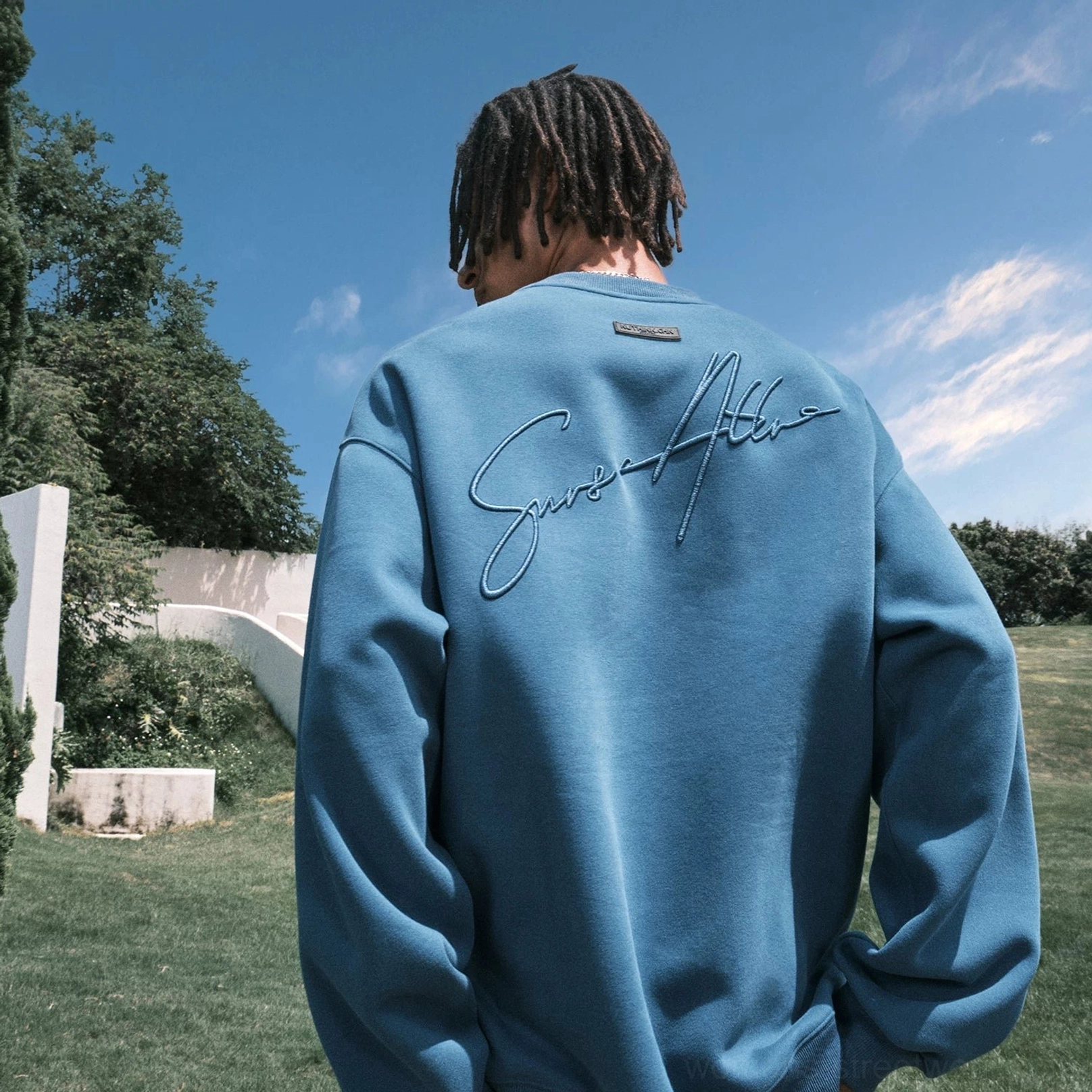 Close rear view of a dusty blue oversized crewneck sweatshirt showing detailed cursive script embroidery, upper-back tab, and relaxed fit