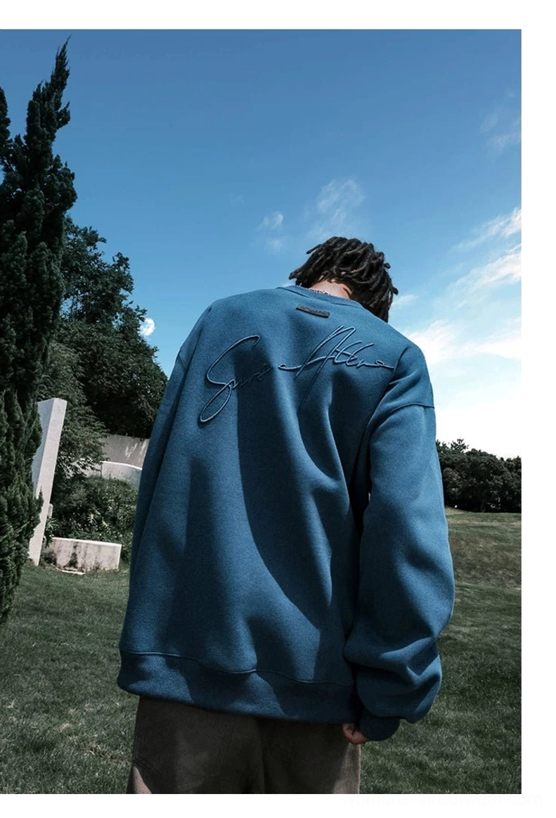 Male model wearing a dusty blue oversized embroidered crewneck sweatshirt with dropped shoulders and brown casual trousers outdoors