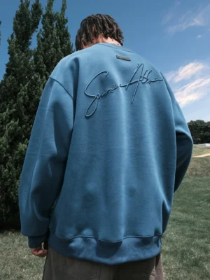 Dusty blue oversized streetwear sweatshirt shown from the back with cursive script embroidery, relaxed sleeves, and a dropped shoulder fit