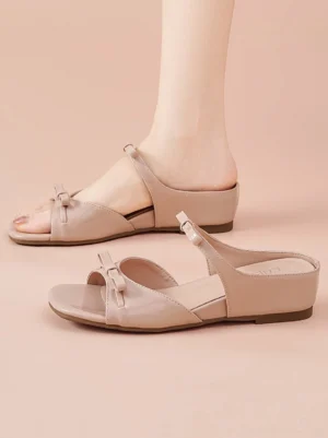 Woman wearing dusty pink bow wedge slide sandals with open square toe, glossy straps, and low wedge heel in a studio setting