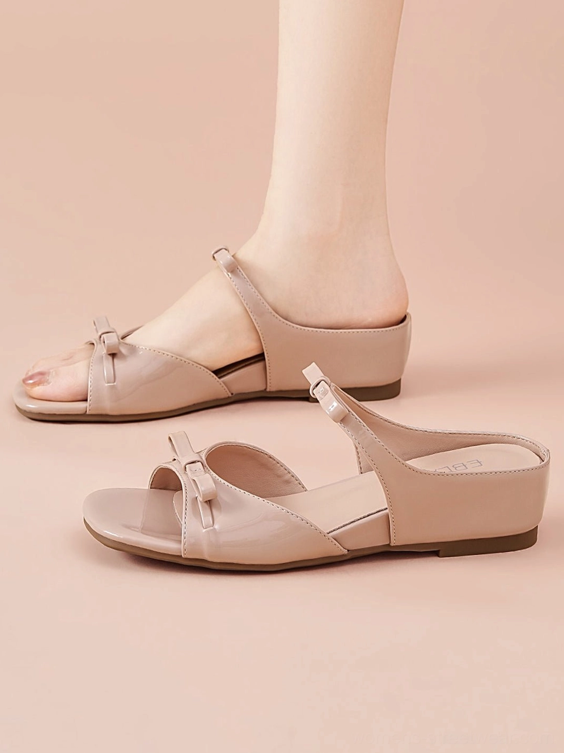 Woman wearing dusty pink bow wedge slide sandals with open square toe, glossy straps, and low wedge heel in a studio setting