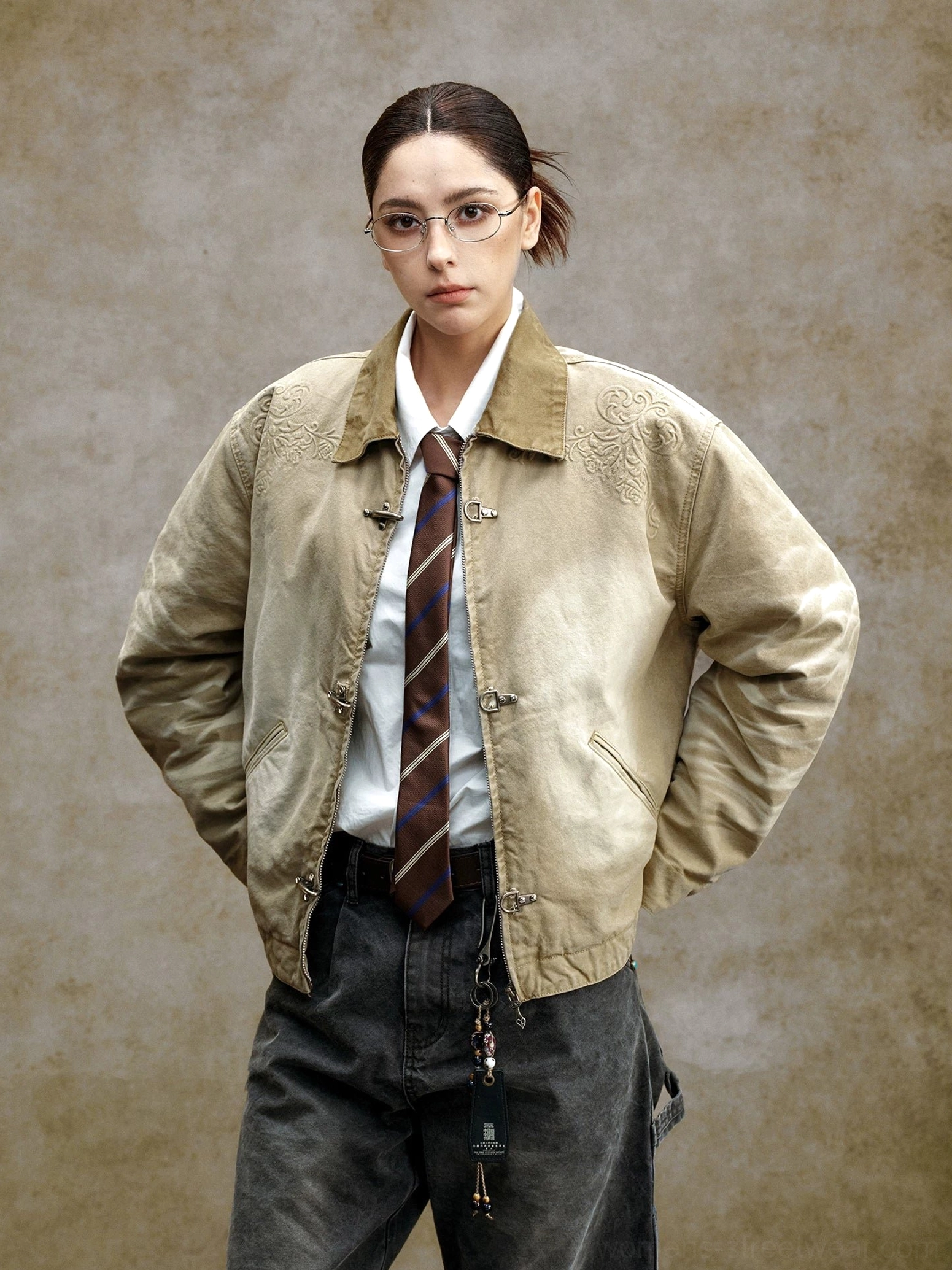 Female model wearing a faded khaki embroidered toggle front utility jacket with contrast collar, formal shirt, tie, and dark workwear trousers