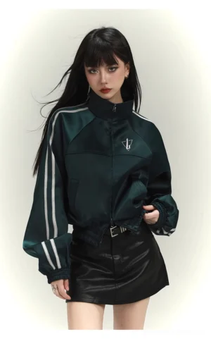 Emerald green cropped satin track jacket worn with a black leather mini skirt, white sleeve stripes, stand collar, and chest V-shaped logo