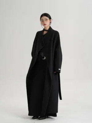 Extra long dark gray wool blend wrap coat shown from the front with an asymmetric wide lapel, relaxed drop shoulder silhouette, long waist belt, wide leg trousers, and pointed leather shoes