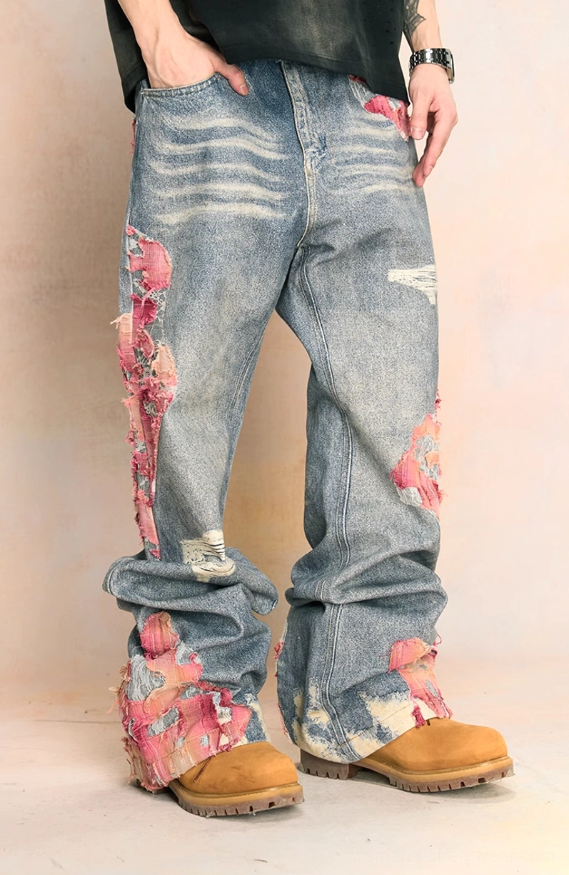 Front view of a male model wearing faded blue wide-leg distressed jeans with pink frayed embroidery patches, black cropped top, and tan boots