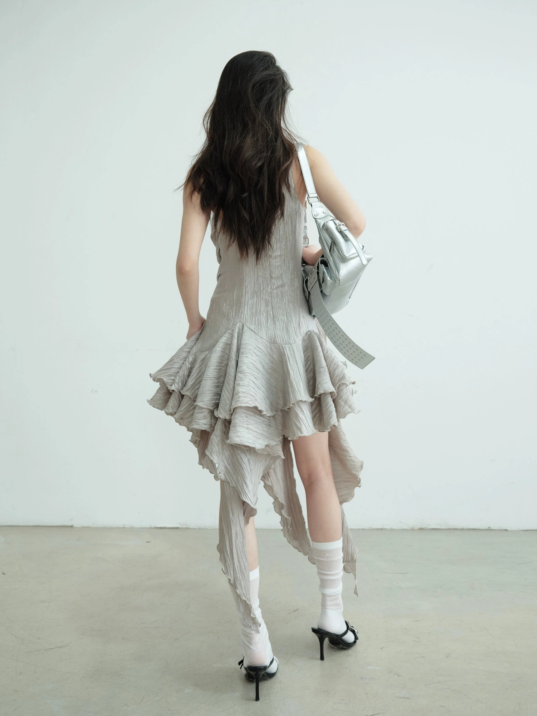 Full back view of a fitted asymmetric metallic ruffle hem party dress with layered high-low hem and silver shoulder bag