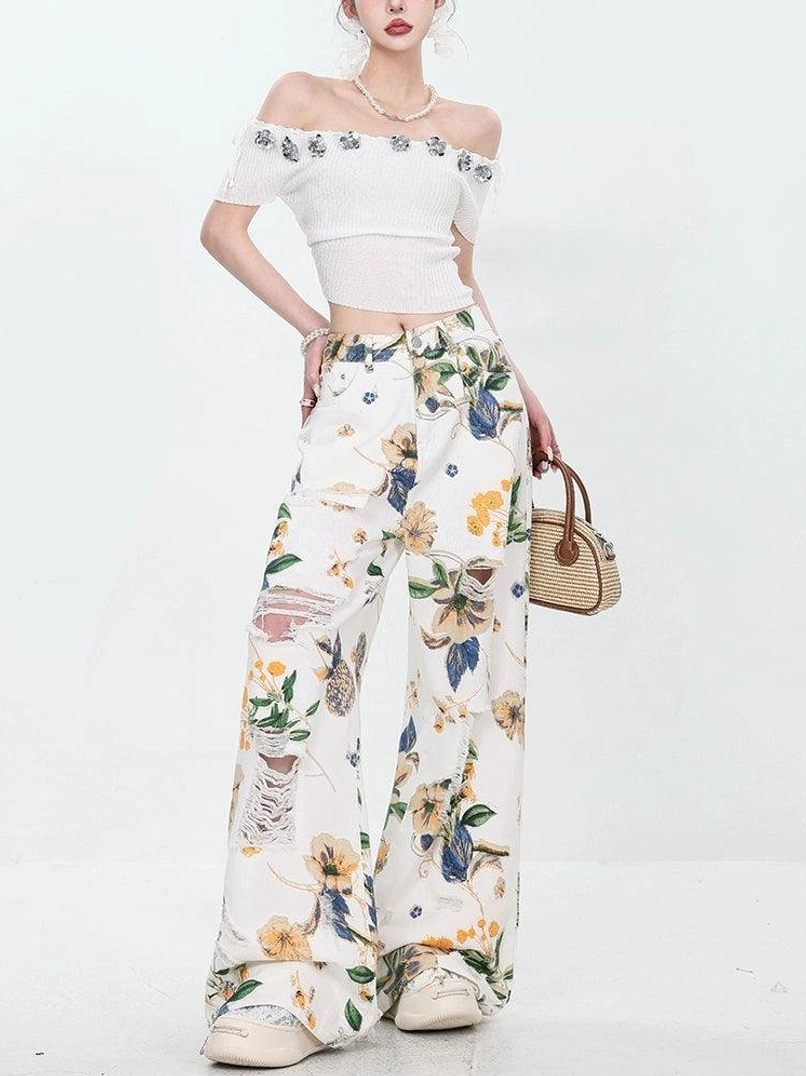 Front three-quarter view of women's floral distressed wide-leg jeans styled with a woven handbag and platform sneakers