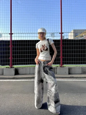 Model wearing washed gray flying bird print convertible wide-leg jeans with vintage portrait artwork, white cropped tee, and cap in an outdoor streetwear setting