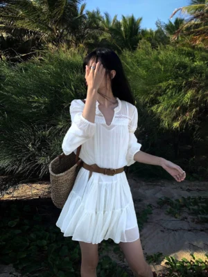 White French ruffle mini shirt dress with V-neck trim, lantern sleeves, pleated hem, brown waist belt and straw tote in a tropical outdoor setting