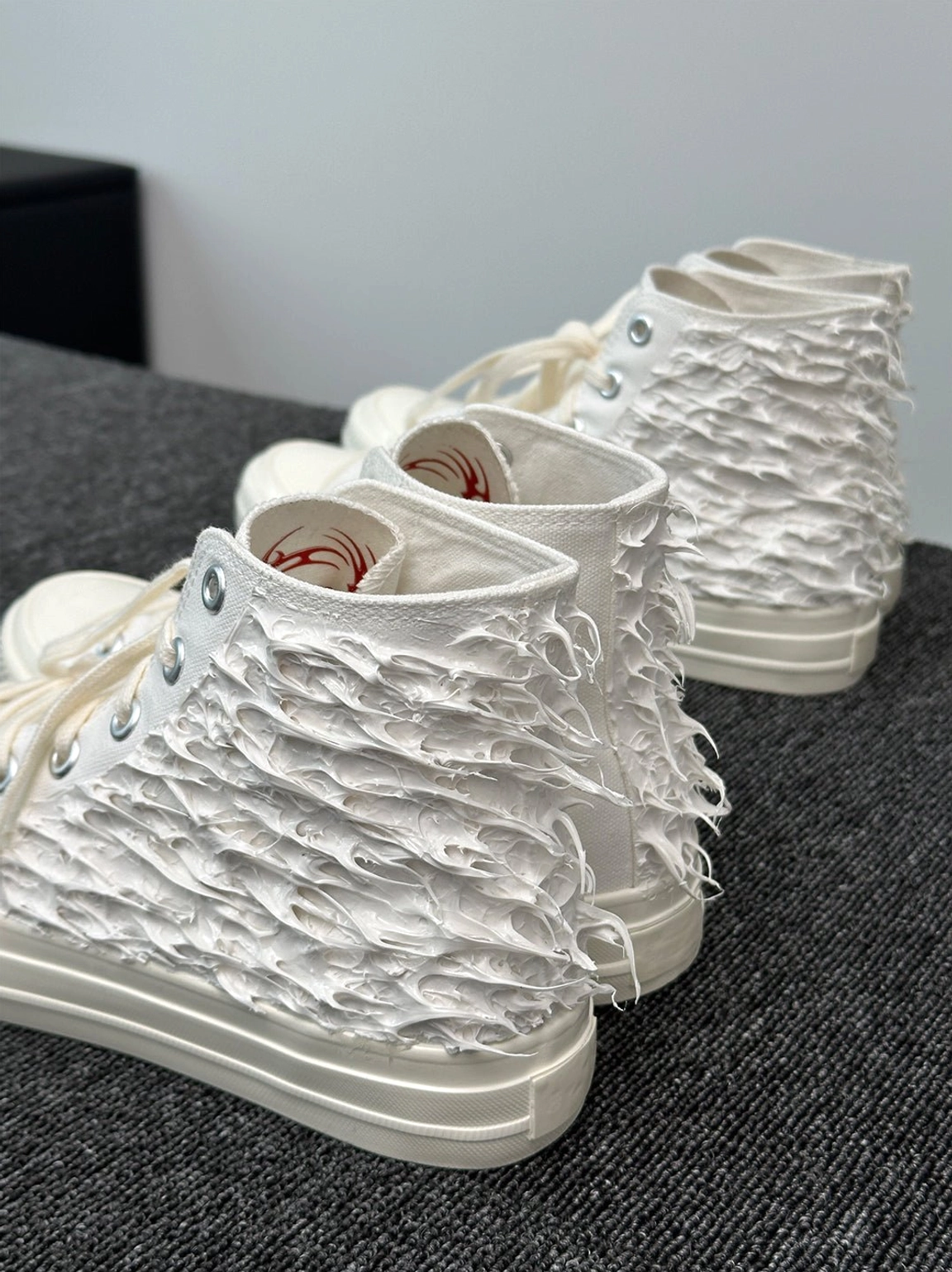 Multiple white fringed high-top canvas sneakers in side flat lay with tassel texture and red tribal print accents