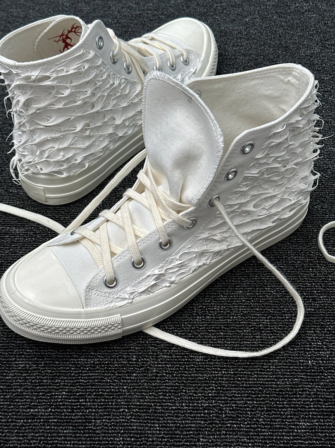 Close-up of fringed white high-top canvas sneaker showing folded tongue, red tribal print and rubber toe cap
