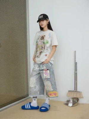 Front view of wide-leg light blue acid-washed denim Bermuda shorts with graffiti artwork, distressed patchwork, heart detail, raw hem, casual tee, baseball cap, and blue slides