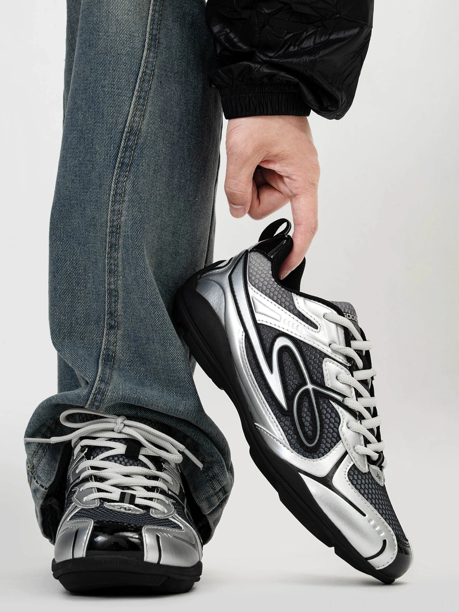 Dark silver futuristic retro runner sneakers held above loose blue denim with metallic overlays black breathable mesh curved wave side detail and lace-up closure
