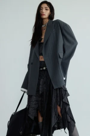Gray genderless striped oversized blazer shown from the front three-quarter view with dark camisole ruffled skirt neck chain and black tote bag
