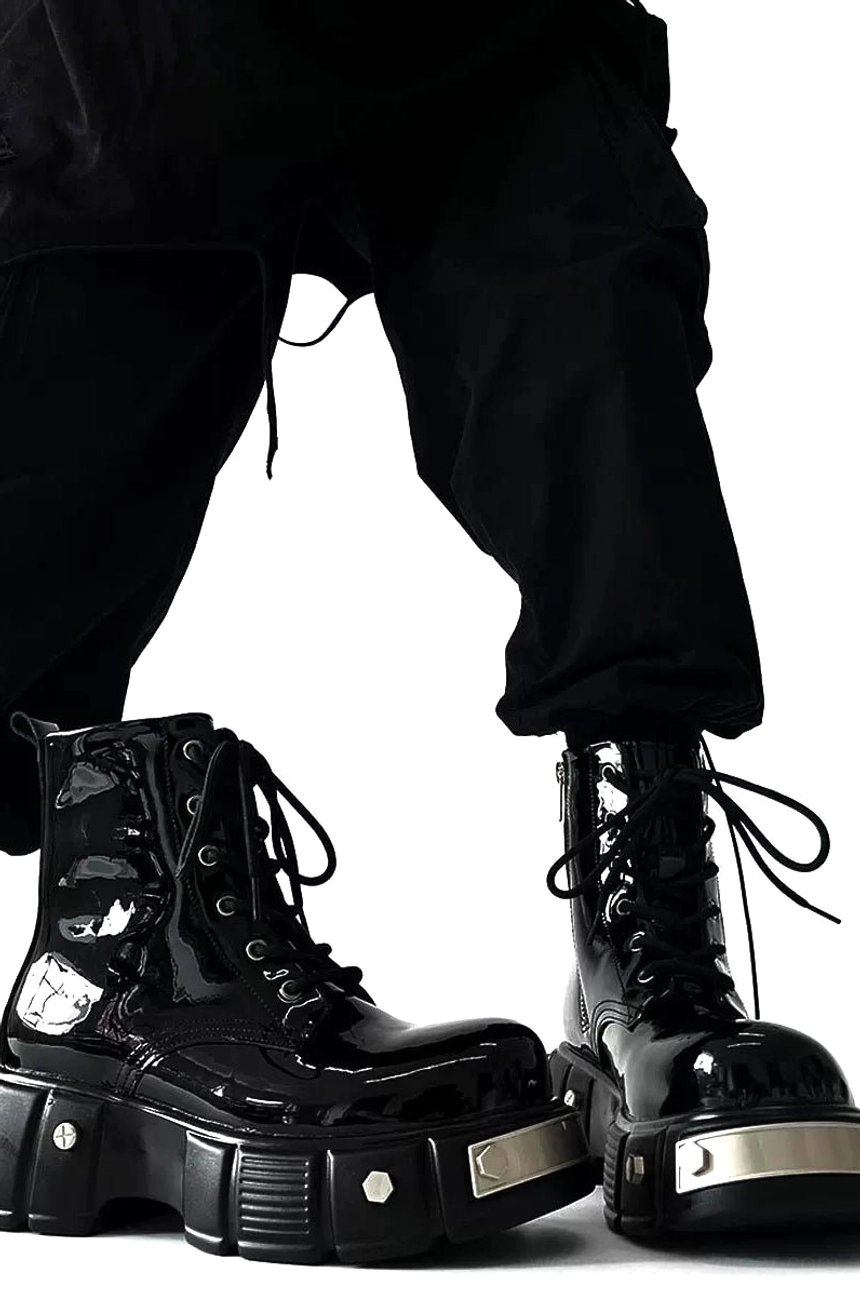 Low-angle front view of glossy black patent leather platform combat boots worn with loose black cargo pants, featuring tied laces, silver front accents, and thick lug soles