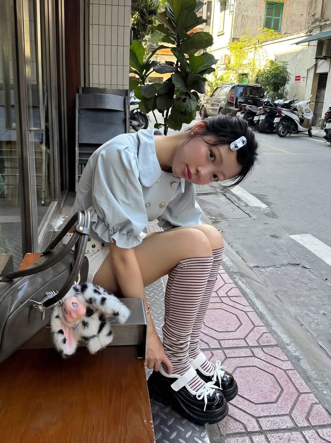 Glossy black and white platform Mary Jane shoes worn with striped over-the-knee socks, white blouse, shorts, and a brown handbag on a city street