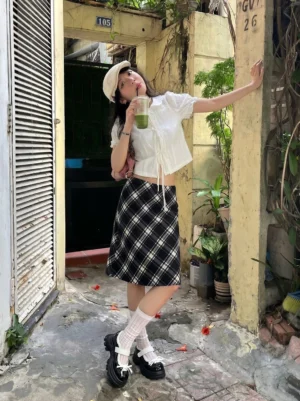 Women's glossy black platform Mary Jane shoes styled with white contrast straps, white knee-high socks, and a black plaid skirt in a vintage streetwear outfit