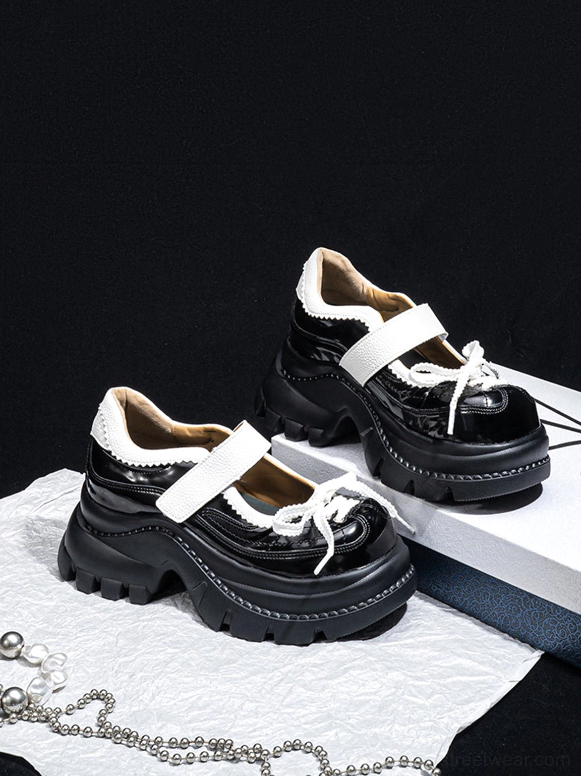 Pair of glossy black and white platform Mary Jane shoes displayed on a black studio background with gift boxes, paper, and pearl chain accents