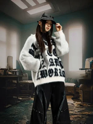 White fuzzy gothic cross sweater shown from the front on a female model with a cat ear bucket hat and black wide leg streetwear pants in an industrial studio