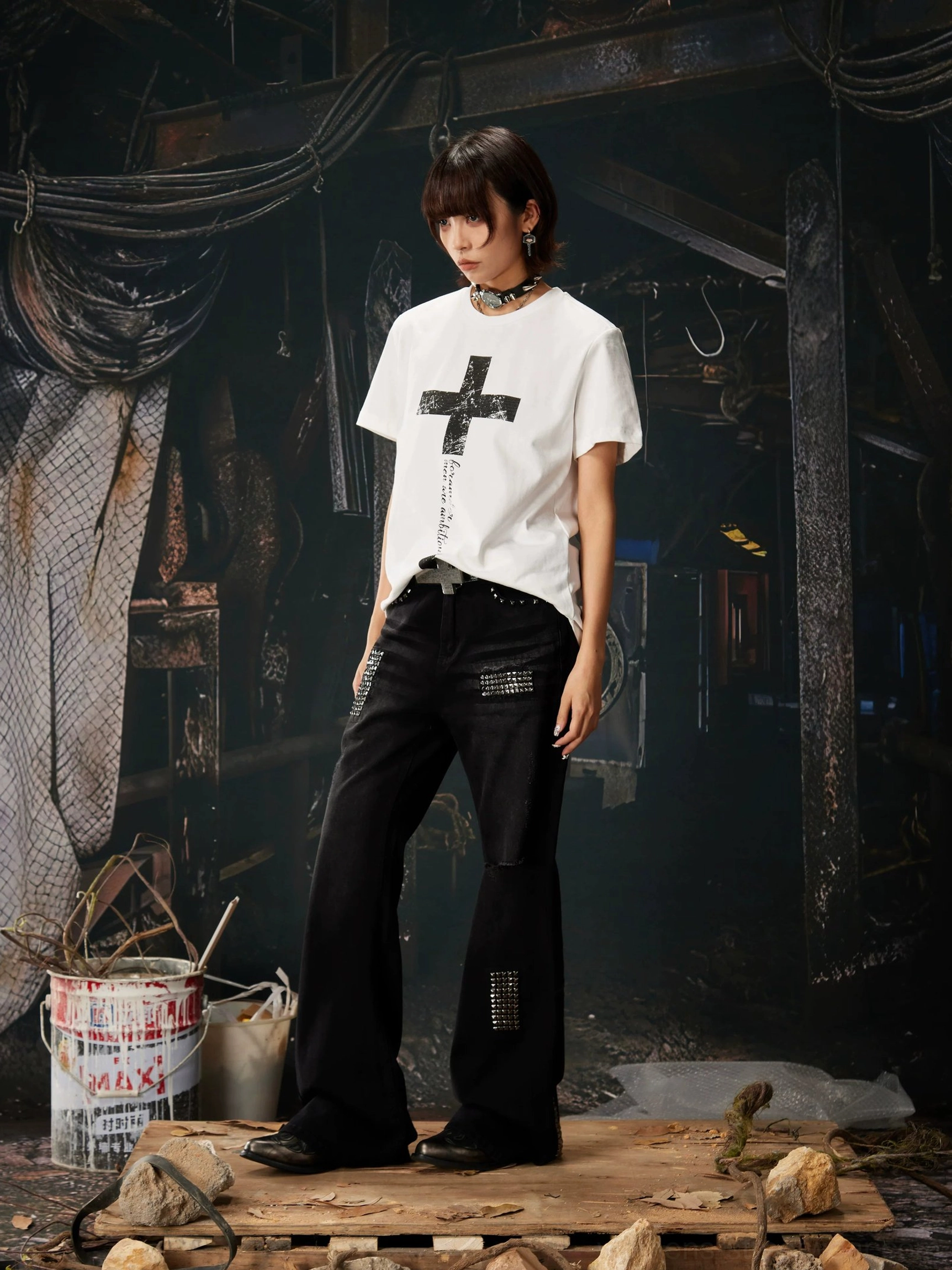 White Gothic cross script graphic streetwear T-shirt side view styled with black flared pants in an industrial setting
