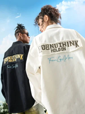 Two models wearing black and off-white graphic embroidered oversized button-up shirts with bold back lettering and handwritten streetwear graphics outdoors